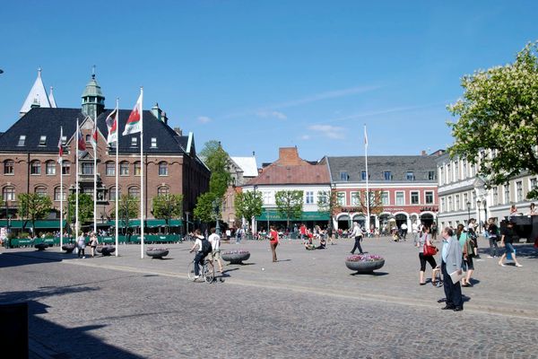 En stadsbild med folk som går, cyklar och samlas i ett öppet torg, omgiven av byggnader med butiker och flaggor, på en solig dag.