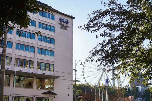 En byggnad med skylt som säger 'World Trade Center Göteborg' och en nöjespark med ett stort pariserhjul och karuseller i bakgrunden, omgiven av träd och blå himmel.